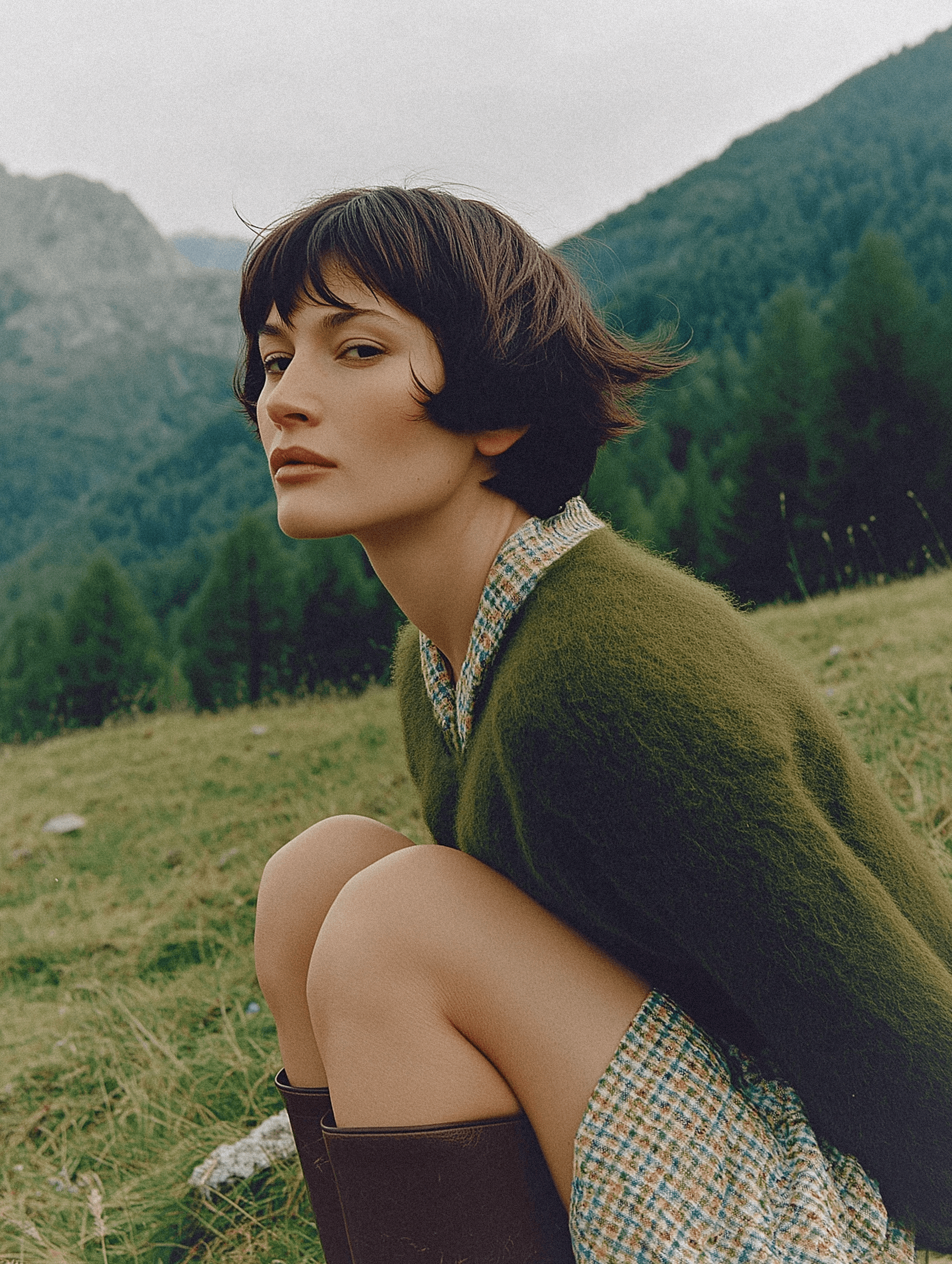 A person with short dark hair crouches in a grassy meadow, wearing a green sweater over a plaid dress and tall boots, set against a backdrop of lush trees and a mountainous landscape.