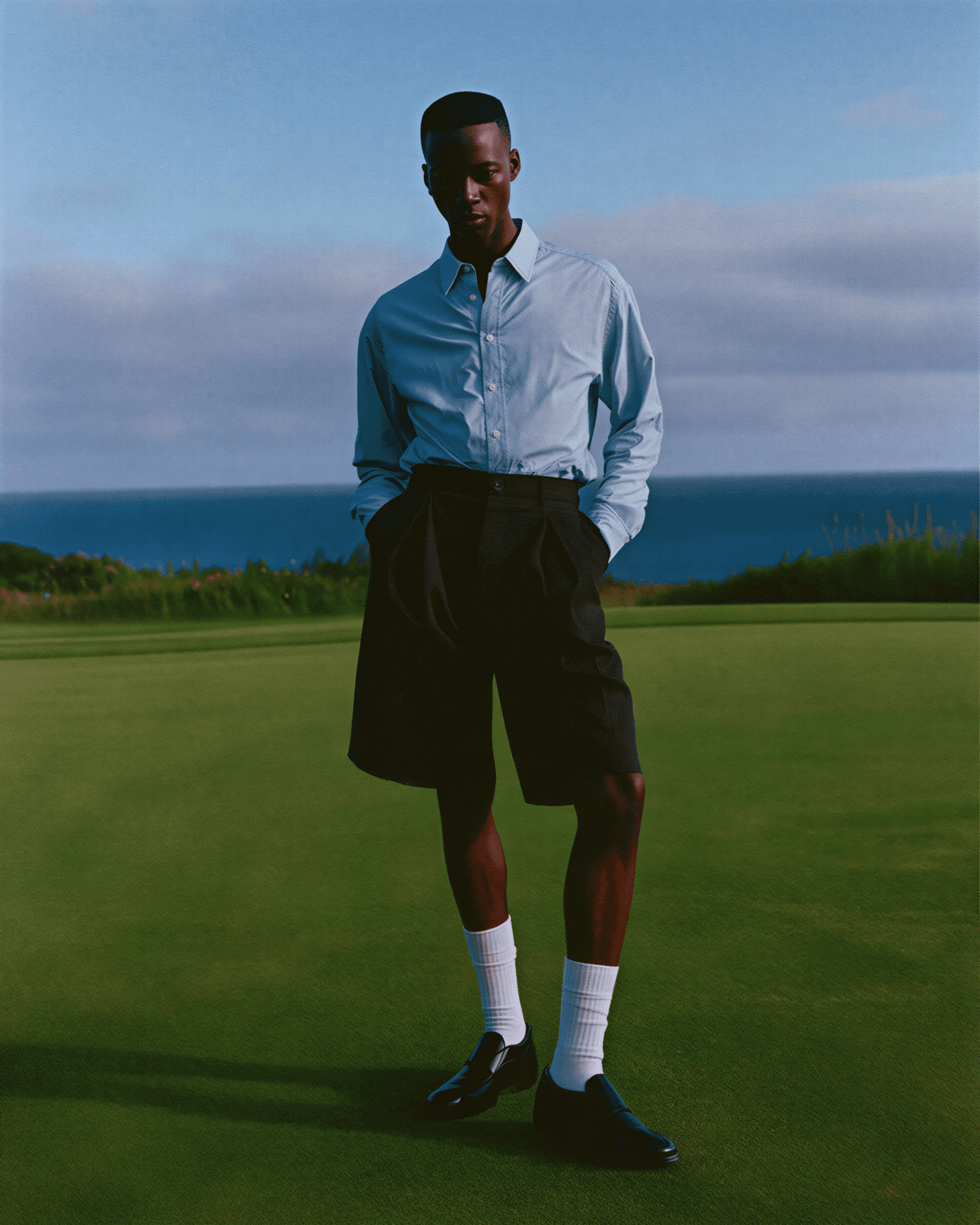 A person stands confidently on a lush green golf course, wearing a light blue shirt, dark shorts, and loafers, with a scenic ocean and sky backdrop.