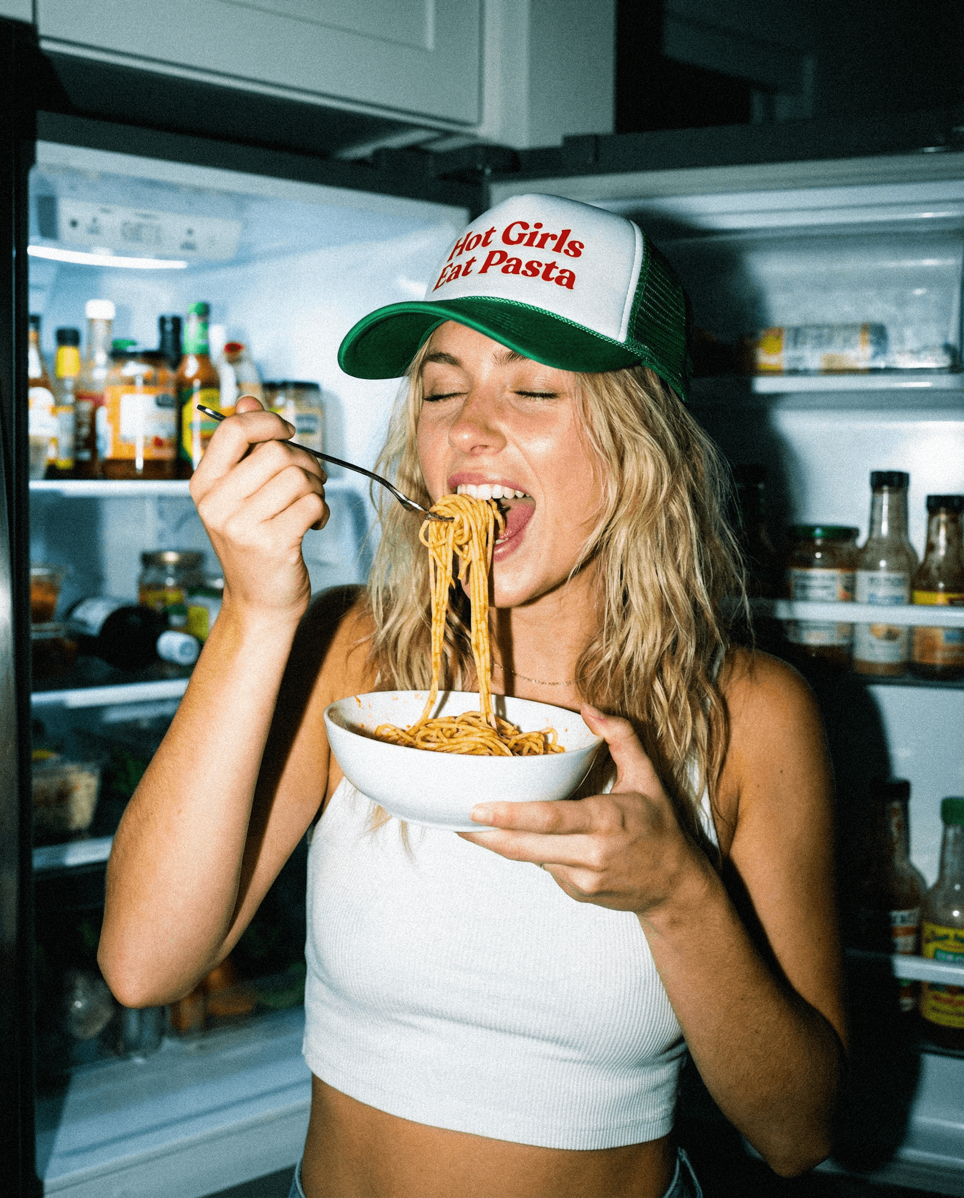 Person eating pasta in a kitchen with an open refrigerator.
