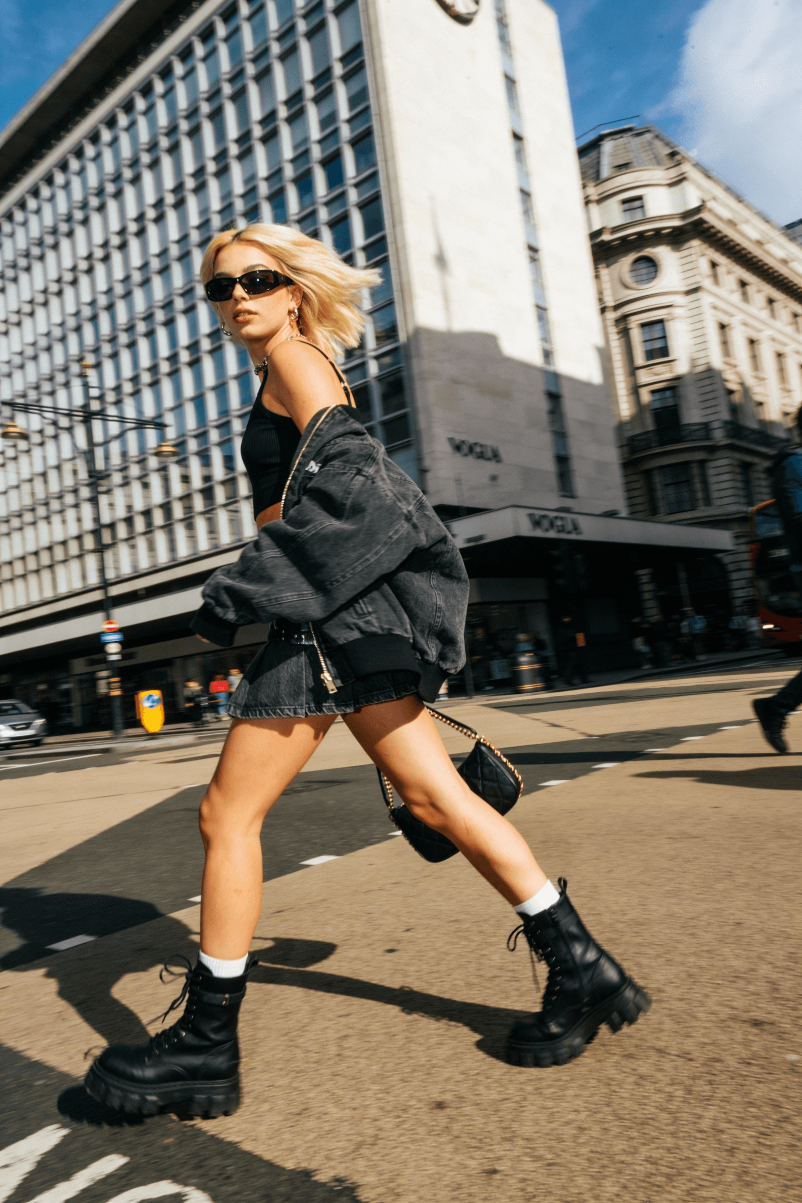 Woman walking on a city street wearing sunglasses, black outfit, and black boots.